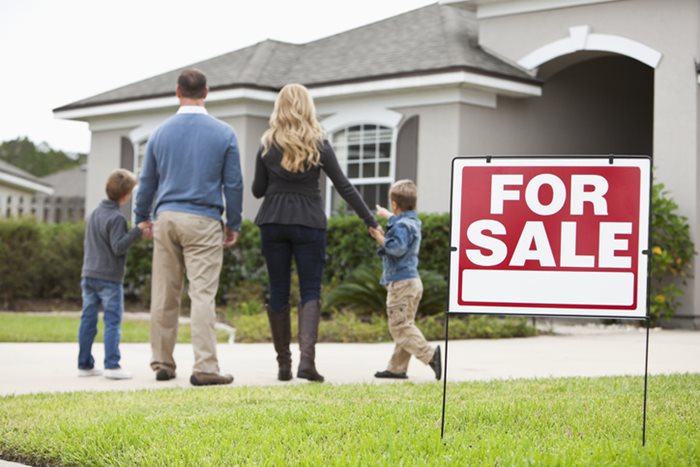 Homebuying with the family, standing in front of a house with a for sale sign.
