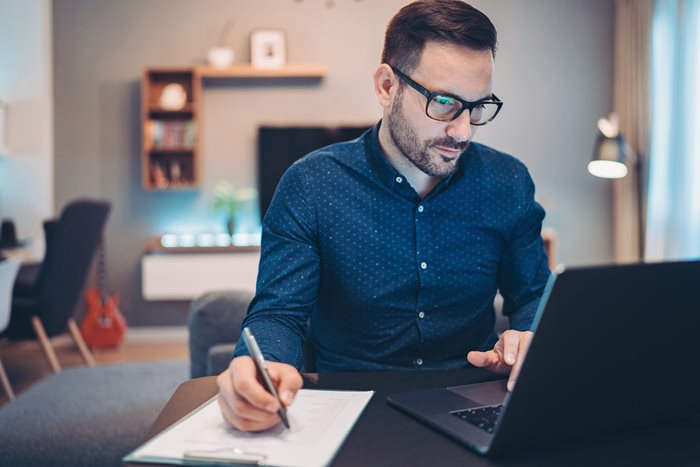 A man looking at his laptop while he takes notes.